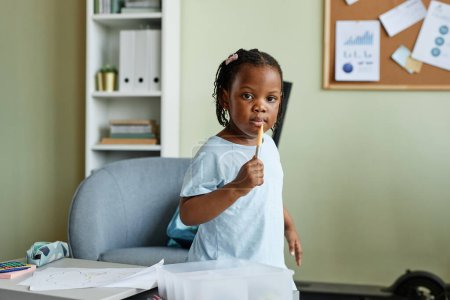 Téléchargez les photos : Portrait de mignonne fille noire tenant le crayon et regardant la caméra tout en dessinant des images au petit bureau - en image libre de droit
