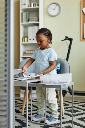Foto de Retrato vertical de longitud completa de la linda chica negra de pie en un pequeño escritorio y dibujos - Imagen libre de derechos