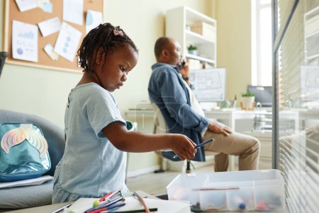 Foto de Retrato de vista lateral de niña negra eligiendo lápices de colores mientras dibuja en la oficina con el padre trabajando en el fondo - Imagen libre de derechos