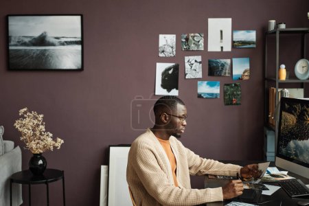 Foto de Hombre negro creativo retocando fotos de paisaje en la computadora en su oficina en casa - Imagen libre de derechos