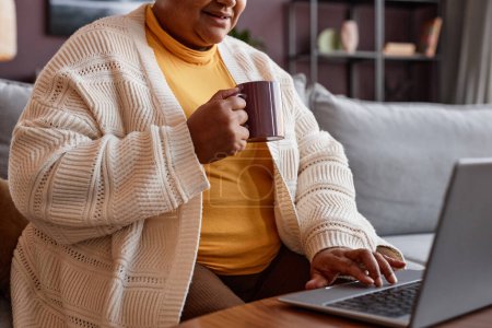 Téléchargez les photos : Plan recadré d'une femme noire âgée utilisant un ordinateur portable à la maison et profitant d'une tasse de café, espace de copie - en image libre de droit