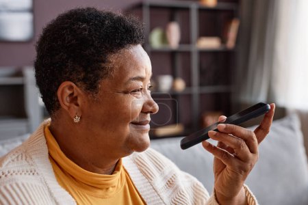 Téléchargez les photos : Portrait latéral d'une femme âgée noire souriante enregistrant un message vocal via un smartphone à la maison, espace de copie - en image libre de droit
