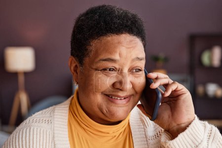 Téléchargez les photos : Gros plan portrait de femme âgée noire appelant par téléphone intelligent dans le cadre de la maison et souriant parlant joyeusement à ses proches - en image libre de droit