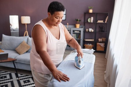 Téléchargez les photos : Vue grand angle de femme noire mature repassage vêtements à la maison tout en prenant soin du ménage - en image libre de droit