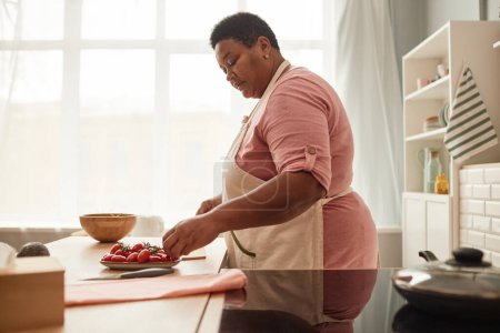 Foto de Vista lateral en la mujer mayor negro usando delantal mientras se cocina en la acogedora cocina casera, espacio de copia - Imagen libre de derechos