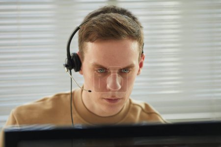 Téléchargez les photos : Portrait d'un jeune homme portant un casque regardant un écran d'ordinateur dans un bureau de soutien technique ou un centre d'appels - en image libre de droit