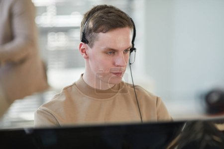 Photo for Portrait of young man wearing headset and looking at computer screen while working in security and surveillance center - Royalty Free Image