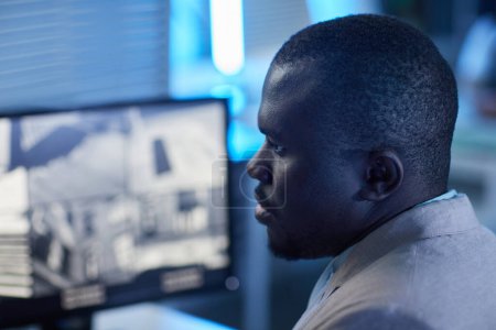 Téléchargez les photos : Portrait en gros plan de l'homme noir regardant les caméras de surveillance dans le centre de surveillance et de sécurité, tons bleus - en image libre de droit