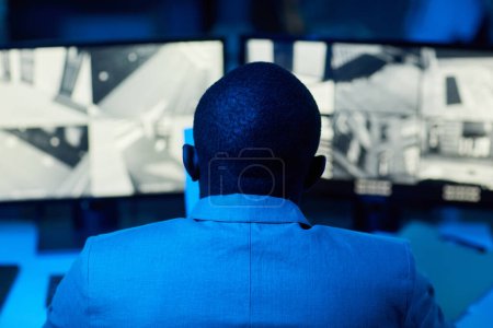 Téléchargez les photos : Vue arrière de l'homme noir chauve regardant les séquences de la caméra de surveillance dans le bureau sombre, les tons bleu et rouge - en image libre de droit