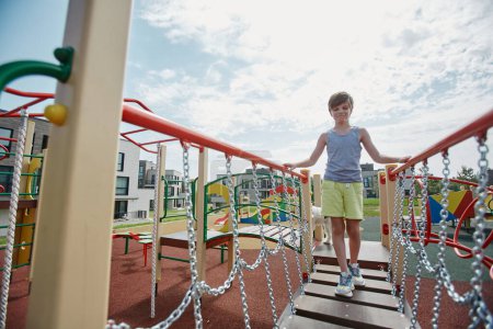 Téléchargez les photos : Portrait complet de jeune garçon jouant sur une aire de jeux colorée et marchant vers la caméra à travers le pont, espace de copie - en image libre de droit
