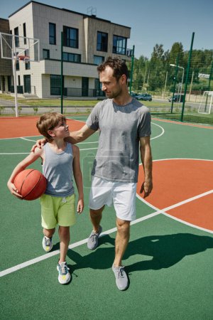 Téléchargez les photos : Portrait vertical complet de père et fils heureux jouant au basket-ball ensemble dans un terrain de sport éclairé par la lumière du soleil - en image libre de droit