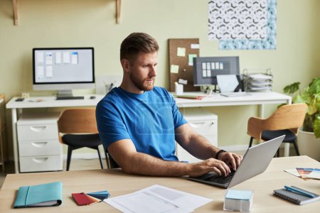 Foto de Retrato de un joven barbudo usando una computadora portátil mientras trabaja en el escritorio en un ambiente acogedor de oficina, espacio para copiar - Imagen libre de derechos