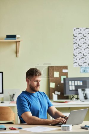Foto de Retrato vertical de un joven barbudo usando una computadora portátil mientras trabaja en el escritorio en un entorno de oficina mínimo - Imagen libre de derechos