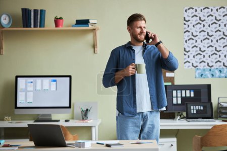 Foto de Retrato de un joven sonriente llamando por teléfono y disfrutando del café mientras toma un descanso del trabajo en la oficina, espacio para copiar - Imagen libre de derechos