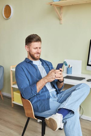 Téléchargez les photos : Portrait vertical d'un jeune homme souriant se relaxant sur le lieu de travail et utilisant un smartphone - en image libre de droit