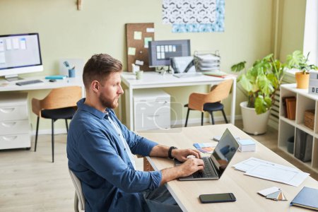 Téléchargez les photos : Portrait latéral de jeune homme barbu utilisant un ordinateur portable tout en travaillant dans un bureau confortable, espace de copie - en image libre de droit