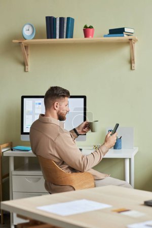 Téléchargez les photos : Portrait vertical de jeune homme utilisant un smartphone tout en prenant une pause sur le lieu de travail dans un bureau minimal - en image libre de droit
