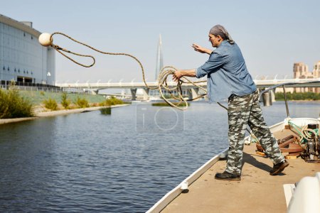 Photo for Full length side view of man standing on boat and throwing line with buoy in water, copy space - Royalty Free Image