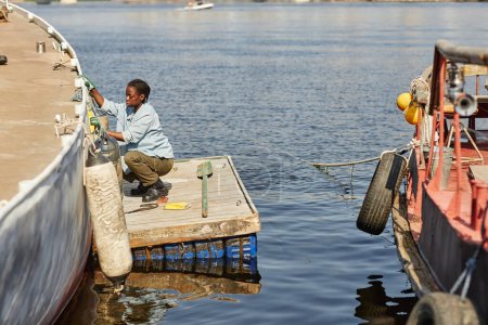 Téléchargez les photos : Portrait grand angle de jeune femme noire nettoyant bateau de pêche dans les quais, éclairé par la lumière du soleil, espace de copie - en image libre de droit