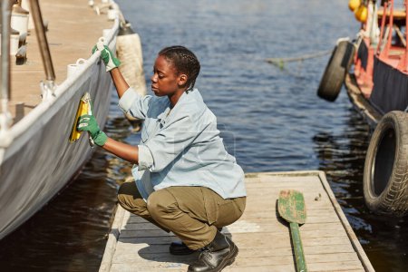 Téléchargez les photos : Pleine longueur vue latérale de jeune femme noire réparer bateau dans les quais éclairés par la lumière du soleil, l'espace de copie - en image libre de droit