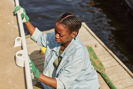 Téléchargez les photos : Portrait grand angle de jeune femme noire nettoyant bateau dans le quai éclairé par la lumière du soleil - en image libre de droit