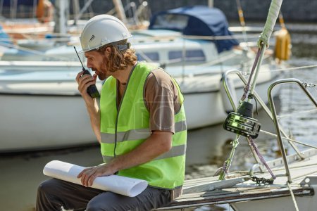 Téléchargez les photos : Portrait latéral du travailleur parlant à la radio dans les quais de yacht éclairés par la lumière du soleil, espace de copie - en image libre de droit