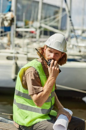 Téléchargez les photos : Portrait vertical de jeune travailleur parlant à la radio dans les quais de yacht éclairés par la lumière du soleil - en image libre de droit
