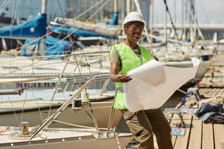 Téléchargez les photos : Portrait de jeune femme noire regardant les plans tout en travaillant dans les quais de yacht éclairés par la lumière du soleil, l'espace de copie - en image libre de droit