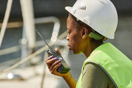 Téléchargez les photos : Portrait latéral d'une jeune femme noire parlant à la radio et portant un casque de sécurité pendant qu'elle travaillait sur un quai - en image libre de droit