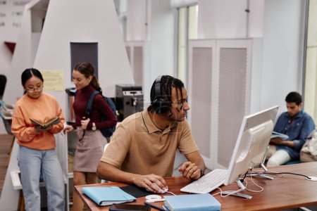 Foto de Retrato de vista lateral de un joven negro usando computadora en la biblioteca universitaria y usando auriculares con gente de fondo - Imagen libre de derechos