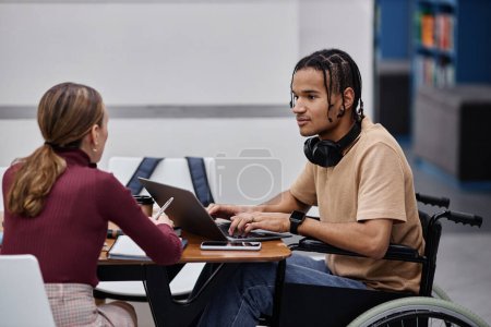 Téléchargez les photos : Vue de côté portrait de jeune garçon noir avec handicap étudiant à la bibliothèque du collège avec un ami - en image libre de droit