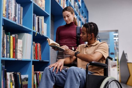 Photo for Portrait of black young man using wheelchair in library and choosing books with friend helping - Royalty Free Image