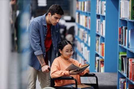 Téléchargez les photos : Portrait de fille asiatique handicapée dans la bibliothèque choisir des livres avec l'aide d'un ami, concept d'inclusion - en image libre de droit