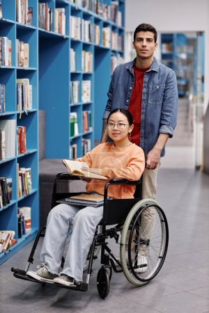 Foto de Retrato completo de un joven sonriente ayudando a una estudiante con discapacidad en la biblioteca mirando a la cámara - Imagen libre de derechos