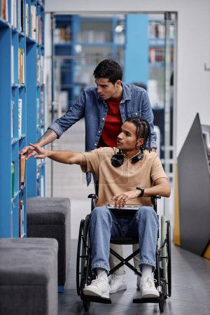 Téléchargez les photos : Portrait vertical complet d'un jeune homme assistant un étudiant handicapé à la bibliothèque et choisissant des livres ensemble - en image libre de droit