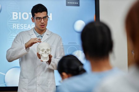 Téléchargez les photos : Portrait à la taille d'un médecin tenant un modèle de crâne lors d'une conférence ou d'un séminaire à l'université de médecine, expliquant la structure osseuse et les méthodes de chirurgie cérébrale - en image libre de droit