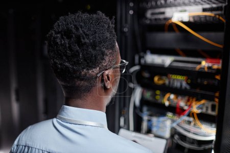 Téléchargez les photos : Vue arrière de l'homme noir en tant qu'ingénieur réseau travaillant avec l'armoire de serveur dans le centre de données, espace de copie - en image libre de droit