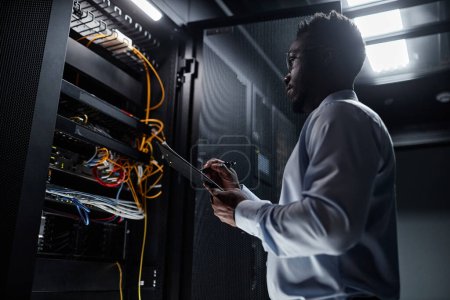 Photo for Backlit side view portrait of black man working with server cabinet in data center and taking notes on clipboard, copy space - Royalty Free Image