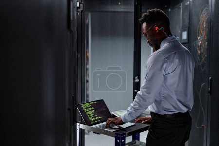 Photo for Side view portrait of black man as network engineer using laptop while setting up servers in data center - Royalty Free Image