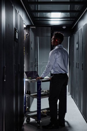 Téléchargez les photos : Portrait vertical complet de l'homme noir en tant qu'ingénieur réseau utilisant un ordinateur portable et configurant des serveurs dans un centre de données - en image libre de droit