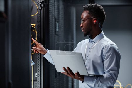 Photo for Side view portrait of young black man as network engineer working with servers in data center and holding laptop - Royalty Free Image