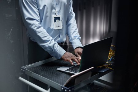 Téléchargez les photos : Gros plan de l'homme noir tapant au clavier de l'ordinateur portable lors de la configuration du réseau dans la salle des serveurs, copier l'espace - en image libre de droit