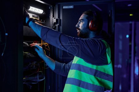 Photo for Side view portrait of bearded man as network technician connecting cables and wires while repairing server in data center lit by neon light - Royalty Free Image