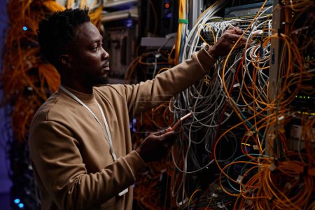 Téléchargez les photos : Vue de côté portrait de jeune homme noir en tant qu'administrateur système serveur de fixation avec des câbles et des fils en désordre - en image libre de droit