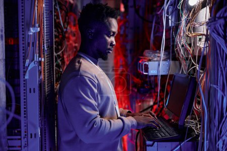 Photo for Side view portrait of black man as network administrator setting up servers and using laptop in neon light - Royalty Free Image