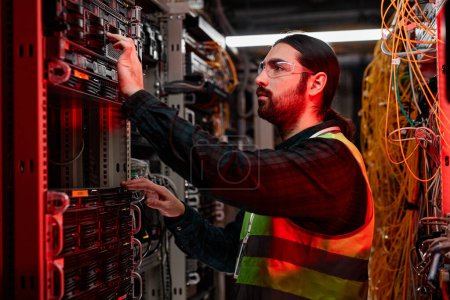 Photo for Side view portrait of bearded network technician inspecting servers and doing maintenance work in data center lit by red neon light - Royalty Free Image