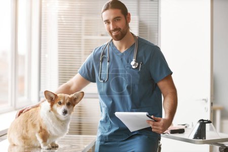 Photo for Young smiling veterinarian with medical document cuddling corgi dog before examination while sitting by workplace in office of vet clinics - Royalty Free Image