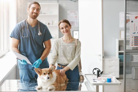 Photo for Happy young owner of corgi dog and male veterinarian in uniform looking at camera while standing by medical table with cute animal - Royalty Free Image
