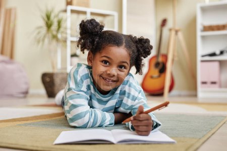Téléchargez les photos : Vue de face portrait de mignonne fille noire allongée sur le sol à la maison et souriant à la caméra écrivant joyeusement dans un carnet ou un journal - en image libre de droit