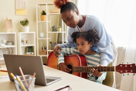 Photo for Portrait of black little girl watching online music lesson with mother and playing guitar at home - Royalty Free Image